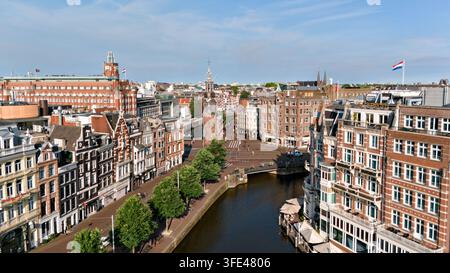 Vista elevata lungo un canale verso la torre Munt, incorniciata da edifici storici sul lungofiume e strade fiancheggiate da alberi nel centro di Amsterdam, Paesi Bassi. Foto Stock