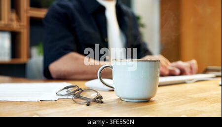 Caffè, scrivania e occhiali in ufficio con un dipendente che digita sulla tastiera per un rapporto o una ricerca. Tazza, documenti e occhiali di una persona al tavolo sul posto di lavoro Foto Stock
