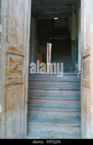 Una porta di legno rustica aperta incornicia la vista di un vecchio corridoio con scala in legno all'interno di un edificio storico. Foto Stock