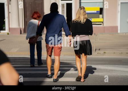 Una coppia attraversa un passaggio pedonale mano nella mano, un altro pedone cammina davanti a loro, le loro lunghe ombre si estendono sulla strada illuminata dal sole Foto Stock
