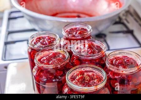 Collezione di barattoli di vetro pieni di vivace marmellata di fragole, che mostra fragole intere e mezze. Una grande pentola con altra marmellata è sfocata in bac Foto Stock