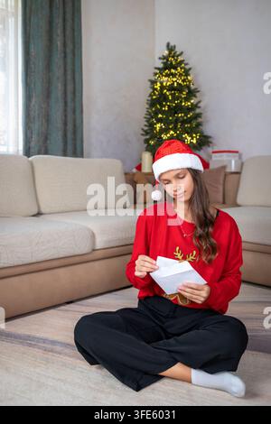 Ragazza con cappello di Babbo Natale che legge una lettera di festa accanto a un albero di Natale illuminato in soggiorno, con ragazza, Babbo Natale, cappello, dettaglio rosso Foto Stock