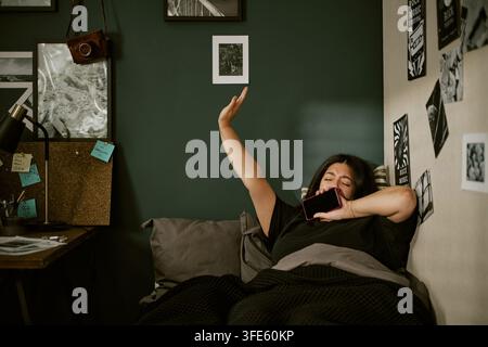 Donna biracial che sbadiglia e si stira con lo smartphone a letto Foto Stock