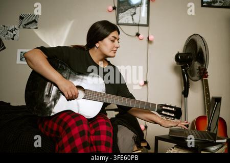 Donna biracial che tiene una chitarra e sfoglia un laptop per la musica Foto Stock