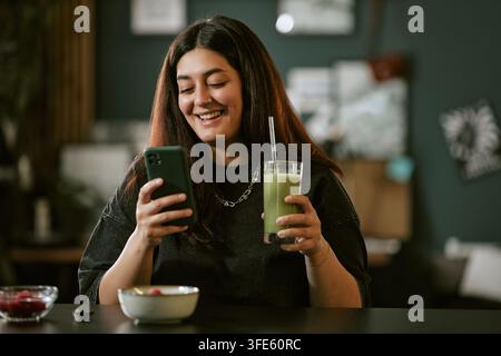 Donna biracial sorridente che usa uno smartphone e tiene un frullato verde Foto Stock