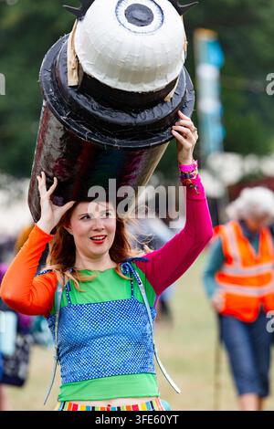 DONNA IN ABBIGLIAMENTO COLORATO PORTA UN PROPS GIGANTE A FORMA DI OCCHIO, REPORTAGE, FESTIVAL GREEN MAN 2018, BRECON, WALES, UK, 2018: Una giovane donna dai capelli rossi e con un trucco labbra brillante, vestita con un abbigliamento colorato a più motivi, sorride mentre bilancia un props gigante fatto a mano a forma di occhio sulla testa durante un evento all'aperto. Foto: Rob Watkins. INFO: Il festival Green Man è iniziato nel 2003 in Galles. Si tiene ogni anno nelle Brecon Beacons il terzo fine settimana di agosto, celebrando musica alternativa, arte, letteratura, scienza e cultura a misura di famiglia in uno splendido scenario montano. Foto Stock