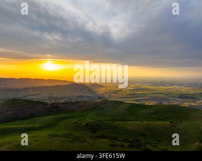 Una veduta aerea cattura un vasto paesaggio sotto un cielo drammatico al tramonto. Colline rigogliose e una piccola città incastonata in una valle creano un'atmosfera serena. Foto Stock