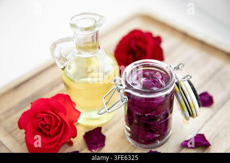 Petali di rosa essiccati in un barattolo coperto con olio per fare olio di rosa a casa. Fiori di rosa di diversi colori disposti in modo decorativo intorno. Foto Stock