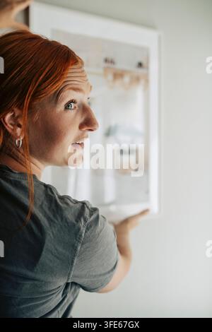 Primo piano di una donna dai capelli rossi che appende un quadro al muro nella sua nuova casa Foto Stock