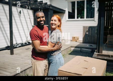 Ritratto di una coppia felice e diversificata che si abbraccia davanti alla loro casa nel giorno del trasloco Foto Stock