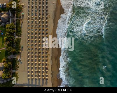Veduta aerea di file di ombrelloni di paglia sulla spiaggia di sabbia con onde turchesi che lambiscono la riva a Chania, Grecia Foto Stock