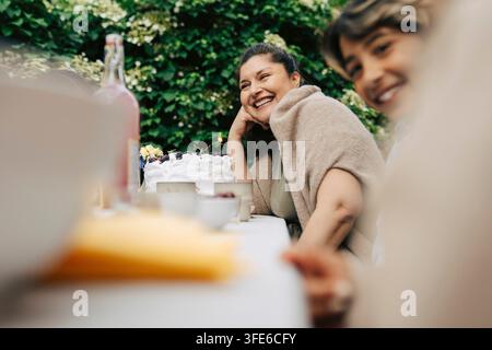 Donna felice appoggiata sul gomito e che ride mentre è seduta al tavolo da pranzo durante una cena nel cortile Foto Stock