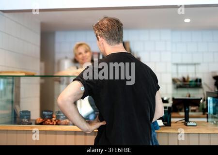 Vista posteriore del proprietario maschile con sensore di glucosio sul braccio che conversa con una collega donna in panetteria Foto Stock