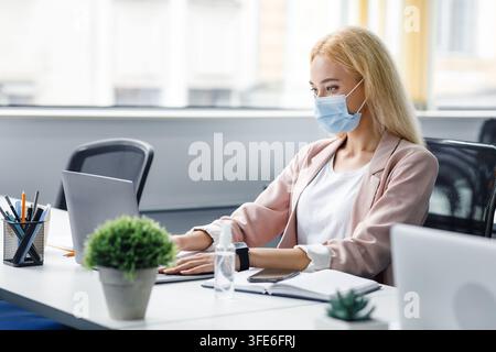 Ufficio moderno protetto da virus e manager al lavoro. Donna d'affari bionda con mascherina protettiva che digita al computer su un tavolo con una bottiglia di disinfettante Foto Stock