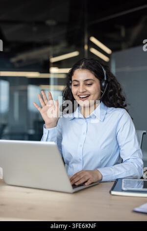 Agente del servizio clienti sorridente con auricolare e mano alzata, lavora su un laptop in un ufficio moderno, fornisce supporto online e comunica con i clienti Foto Stock