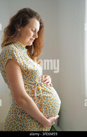 Donna incinta tiene il ventre, lo accarezza con tenerezza soddisfatta e gioiosa al chiuso. Madre in attesa che sente il suo bambino mostra gioia della gravidanza a quarant'anni e protezione materna Foto Stock