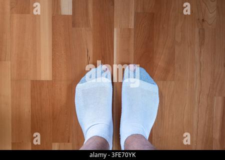 Vista dall'alto di una persona in piedi su un pavimento in parquet chiaro che indossa calzini grigi e bianchi con un buco alle punte. Foto Stock