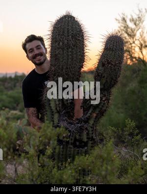 Deserto di Sonora, Arizona - Luglio 2024: un uomo sorridente si trova accanto alle braccia di un grande cactus saguaro silouettato contro un cielo al tramonto. Foto Stock