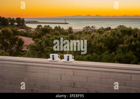 Due tazze di espresso su una ringhiera del balcone contro un vivido sorgere dorato sul mare mediterraneo e sugli alberi costieri. Foto Stock