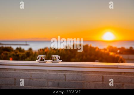 Due tazze di espresso su una ringhiera del balcone contro un vivido sorgere dorato sul mare mediterraneo e sugli alberi costieri. Foto Stock