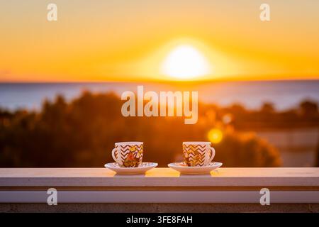 Due tazze di espresso su una ringhiera del balcone contro un vivido sorgere dorato sul mare mediterraneo e sugli alberi costieri. Foto Stock