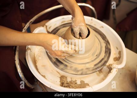 Le mani di Potter modellano una ciotola di argilla su un tornio in una bottega, circondate da strumenti e materiali per la ceramica Foto Stock