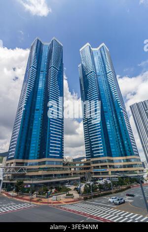 Due grattacieli in vetro blu si ergono sopra un complesso ad uso misto e un ampio incrocio stradale sotto un cielo parzialmente nuvoloso. Foto Stock