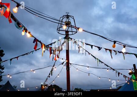LUCI E BANDIERINE DEL FESTIVAL CONTRO UN CIELO SERALE, REPORTAGE, FESTIVAL GREEN MAN 2019, BRECON, GALLES, REGNO UNITO, 2019: Catene di bandiere festive e lampadine luminose sono appese a un palo centrale contro un cielo serale cupo, blu e grigio. Foto: Rob Watkins. INFO: Il Festival Green Man è iniziato nel 2003 in Galles. Si tiene ogni anno nel terzo fine settimana di agosto nelle Brecon Beacons e celebra musica alternativa, arti, letteratura, scienza e cultura a misura di famiglia in uno splendido scenario montano. Foto Stock