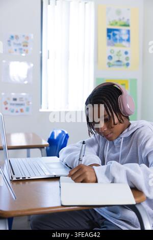 Adolescente africano con felpa con cappuccio, auricolari rosa, scrive su un quaderno in classe usando un laptop, spazio copia Foto Stock