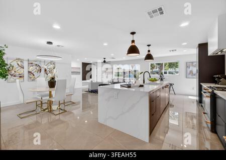 Cucina luminosa e ariosa e sala da pranzo caratterizzata da una grande isola con tetto in marmo, pavimento piastrellato lucido, armadi bianchi, illuminazione a sospensione e un posto a sedere Foto Stock
