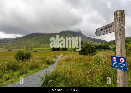 Un cartello indica la strada in una giornata nuvolosa verso Beddgelert con Y Garn e Mynyd Drws Y Coed sullo sfondo Foto Stock