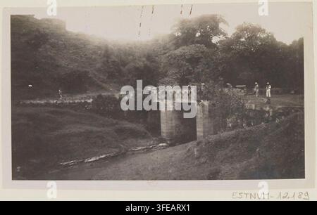 Un ponte di pietra con un unico arco attraversa un canale stretto a Leshan, Sichuan, Cina, circondato da alberi fitti e terreno in pendenza, con diverse figure in piedi sul ponte e nelle vicinanze, di Victor Segalen, un fotografo francese, nel 1914, come parte della Mission archéologique, Chine, 1914. Foto Stock