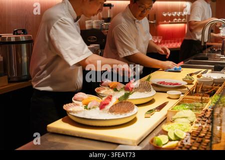 Berlino, Germania - 19 agosto 2025: Due chef maschi asiatici che preparano abilmente sushi con frutti di mare freschi su un bancone di legno, mostrando esperienza culinaria an Foto Stock