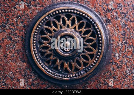 Griglia metallica rotonda ornata montata su un muro di granito rosso. Dettaglio architettonico vintage e concetto di dispositivo di sicurezza classico. Foto Stock