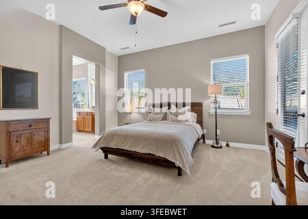 Scena di camera da letto luminosa con biancheria da letto su una struttura in legno, lampade morbide e cassettiere, grandi finestre con tende e ventilatore a soffitto che crea un riposo tranquillo Foto Stock