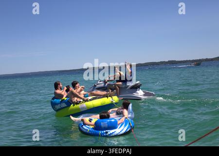 Un gruppo di giovani amici che si godono una giornata estiva soleggiata su un lago blu cristallino, navigando su tubi gonfiabili e un moto d'acqua personale. Foto Stock