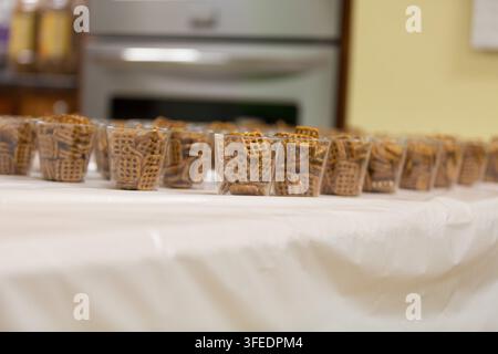 Più bicchieri di plastica trasparenti ripieni di pretzel waffle disposti in file su un tavolo bianco per un bar di snack per feste o un raduno evento. Foto Stock
