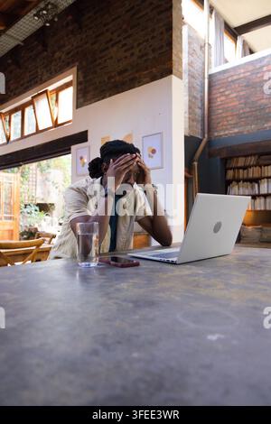 Uomo africano seduto a un tavolo lungo in un spazio di lavoro in mattoni, con la testa appoggiata accanto a un laptop, uno smartphone, acqua Foto Stock