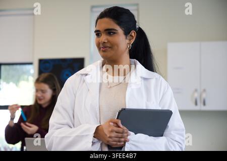 Insegnante indiana in camice da laboratorio bianco, adolescente che maneggia un tablet e una provetta blu nel laboratorio Foto Stock