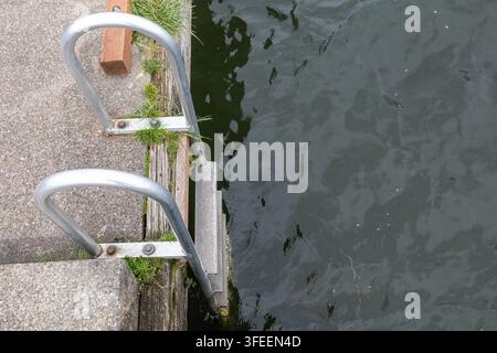 Vista dall'alto di una scala da nuoto in metallo che scende dal bordo di un molo stagionato nell'acqua scura e increspata. Foto Stock