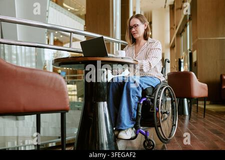 Donna con disabilità che utilizza un laptop in sedia a rotelle al chiuso Foto Stock