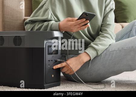 Indipendenza energetica e alimentazione di emergenza, persona che ricarica lo smartphone con una stazione di potenza portatile durante un blackout o una vita fuori rete in una casa moderna Foto Stock