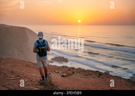 Uomo con zaino che osserva il tramonto sull'oceano a Legzira in Marocco Foto Stock