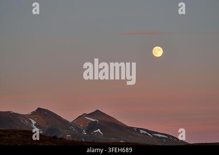 Luna piena che sorge sulle montagne innevate al crepuscolo Foto Stock