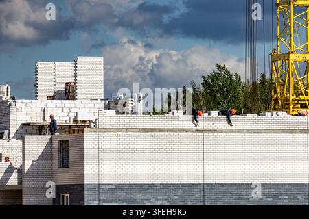 Squadra di costruzione professionale che posa mattoni al piano superiore di un moderno progetto edilizio sotto un cielo sereno e una gru gialla. Foto Stock