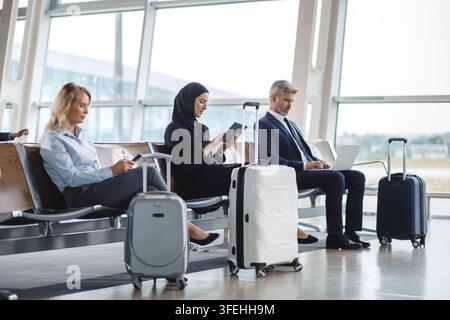 Persone di diverse etnie e generi sono sedute nella sala d'attesa di un aeroporto, ognuna impegnata con i propri dispositivi elettronici e circondata da Foto Stock