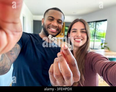 Coppia interrazziale felice che celebra l'acquisto di una nuova casa, tiene in mano una chiave della casa e sorride alla telecamera in un interno moderno, simboleggiando la proprietà Foto Stock