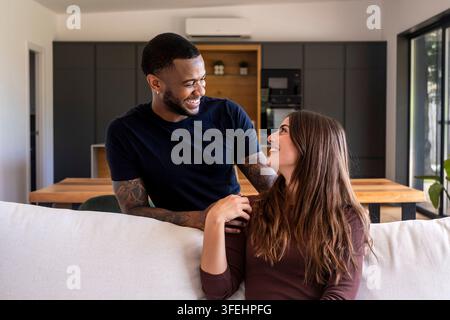 Giovane coppia interrazziale che sorride e ride insieme su un divano in un soggiorno moderno, condividendo un momento caldo di affetto e connessione a casa. Foto Stock