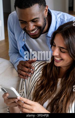Coppia felice che ride insieme mentre guarda uno smartphone a casa, condividendo un momento gioioso di connessione e intimità, partner che godono della tecnologia Foto Stock