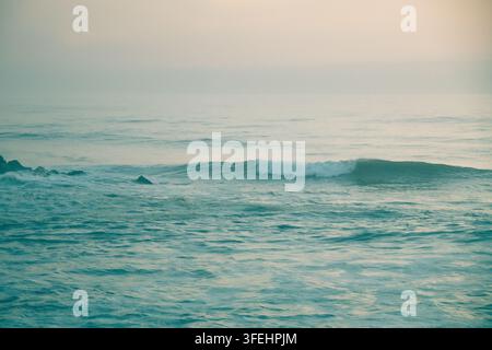 Un paesaggio marino calmo e minimalista che mostra dolci onde oceaniche che si infrangono su scogli scuri sotto un orizzonte morbido e nebbioso al crepuscolo. Pacifico e un Foto Stock